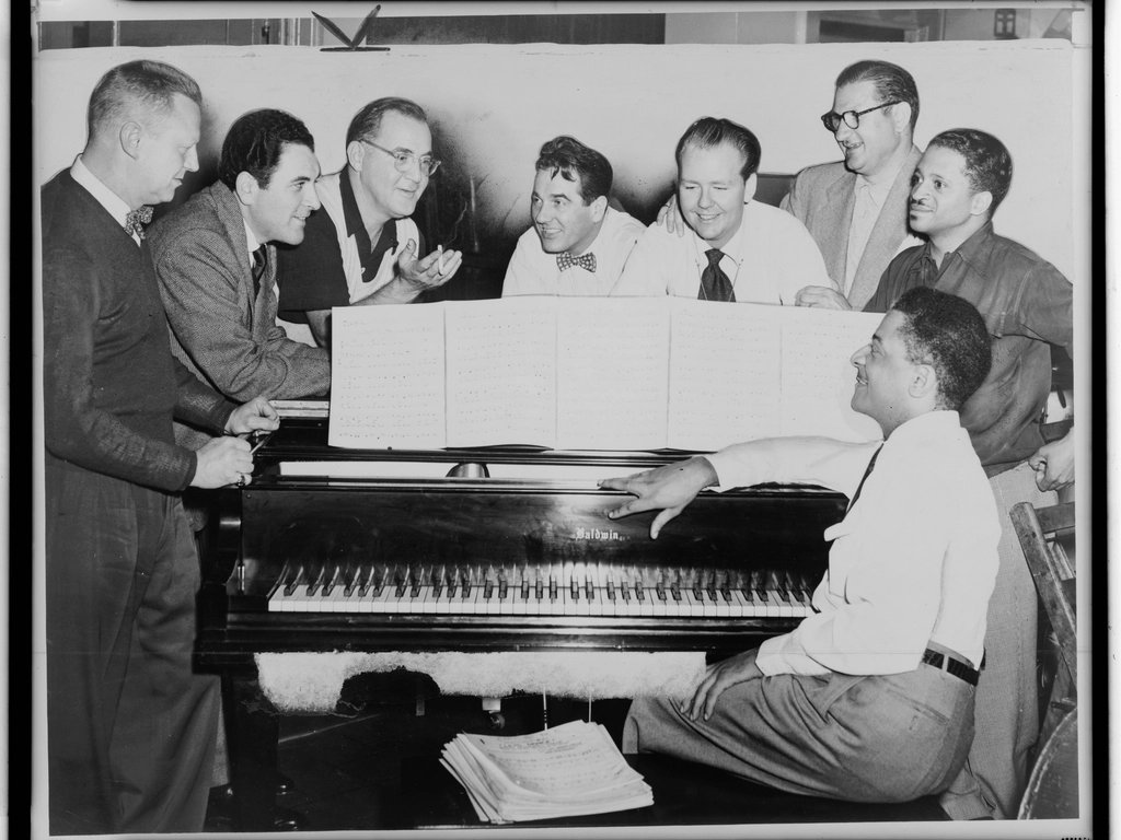 An old photo of a group of musicians round a piano, working on a song together.