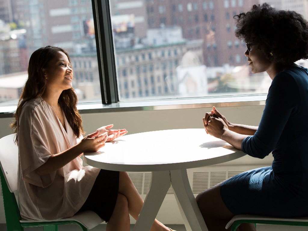 Two women talking across a small round table by a large window.
