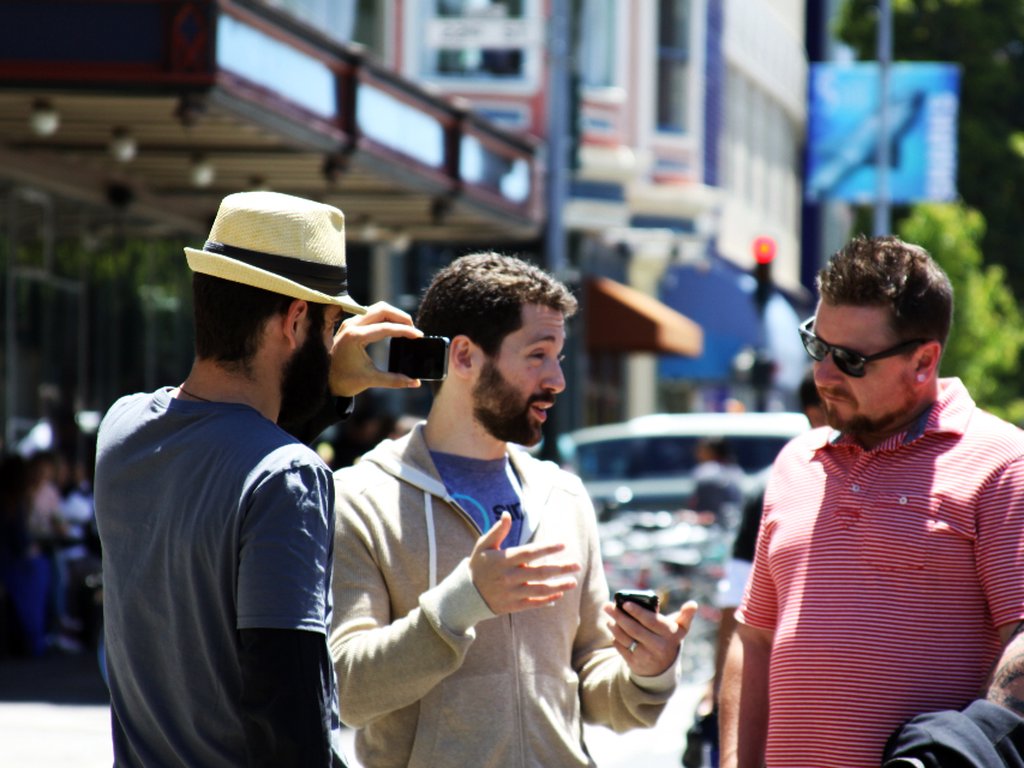 Three men chatting on a busy, sunny street, one of them holding up a phone.