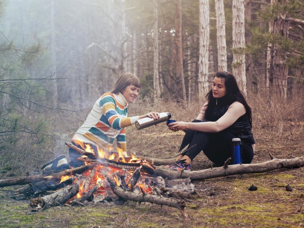 Two women sitting by a campfire in a forest, one passing the other a flask.
