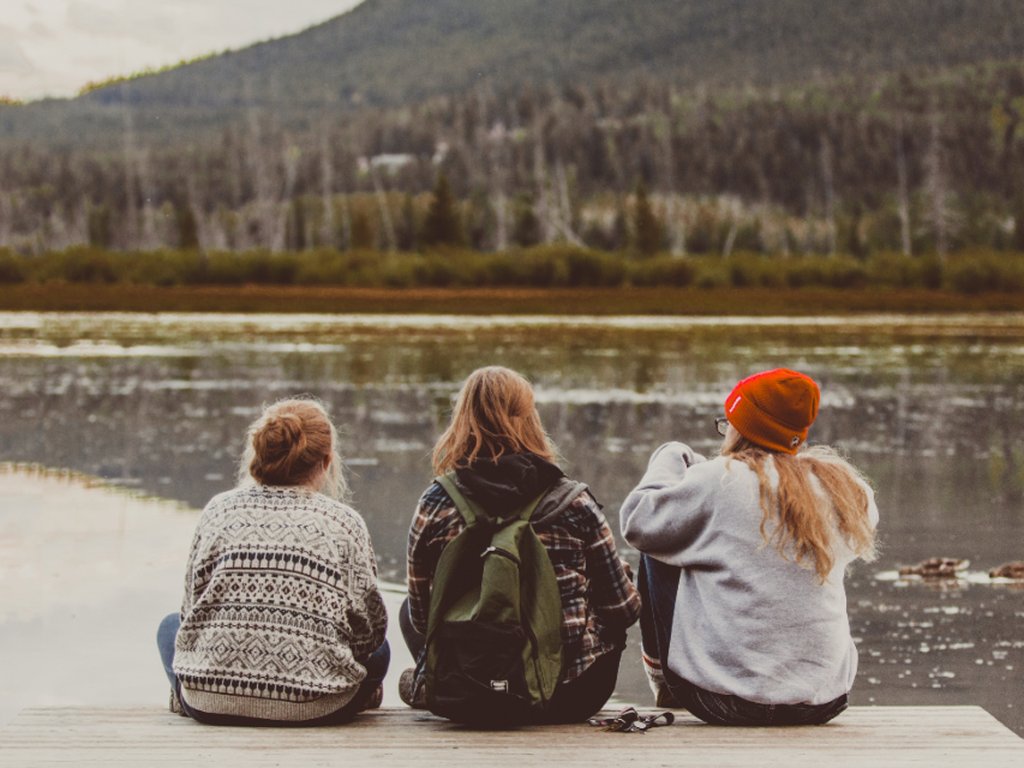 Three people sitting side by side on a jetty by a lake, seen from behind.