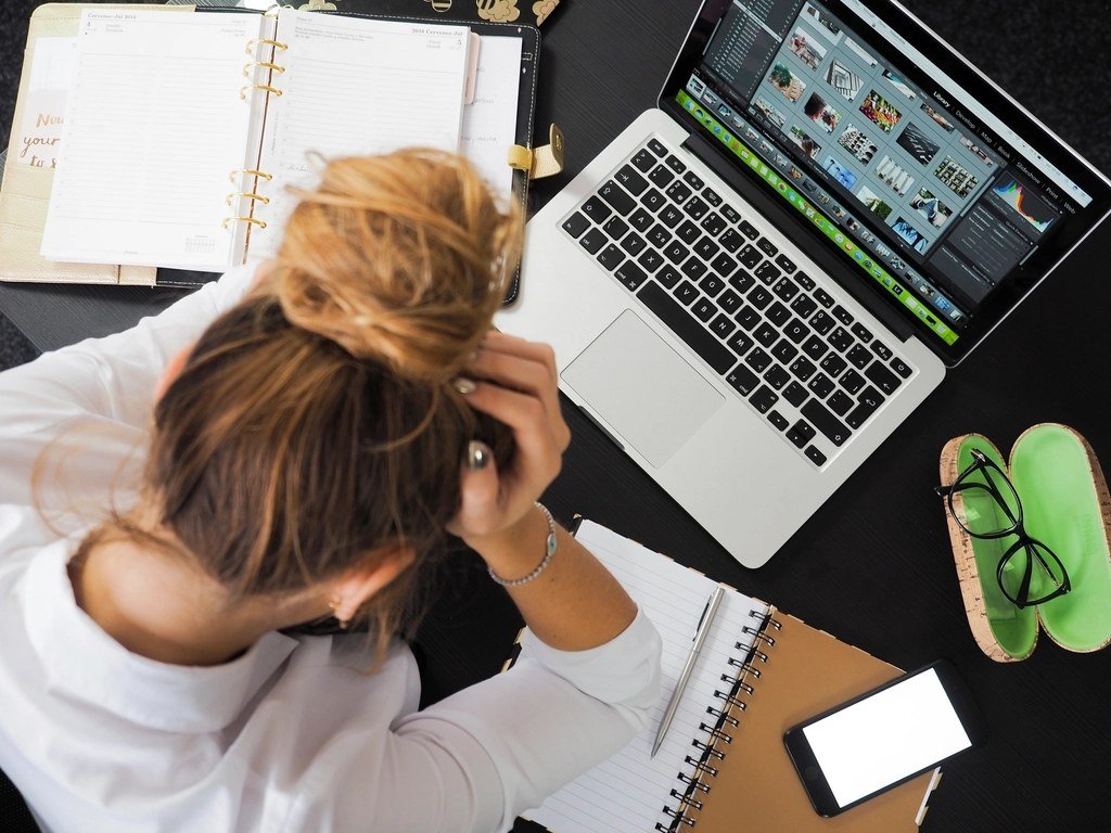 A woman at her desk with her head in her hands, her laptop open and papers all around her.