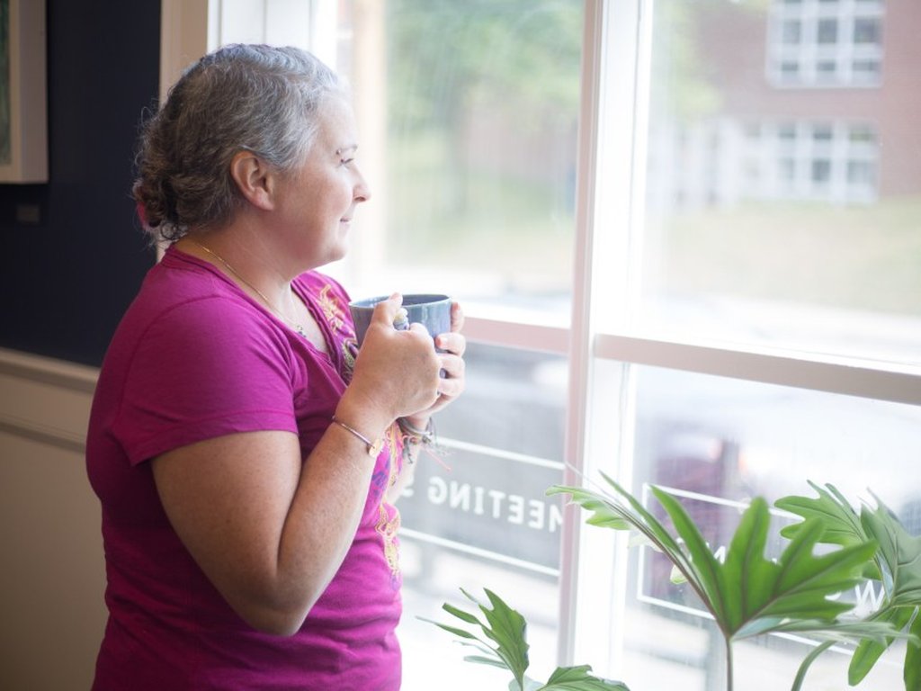 A woman standing by an office window with a mug, taking a quiet moment away from her desk.