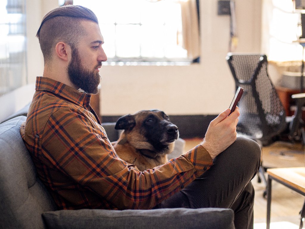 A man on a sofa reading something on his phone, with his dog beside him.