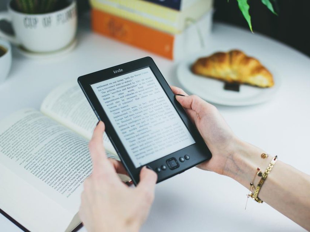 Someone holding an e-reader at a breakfast table, next to an open printed book.