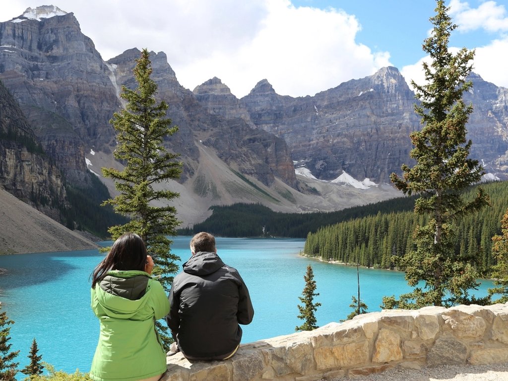 Two visitors looking at a blue lake and high mountains in a national park.