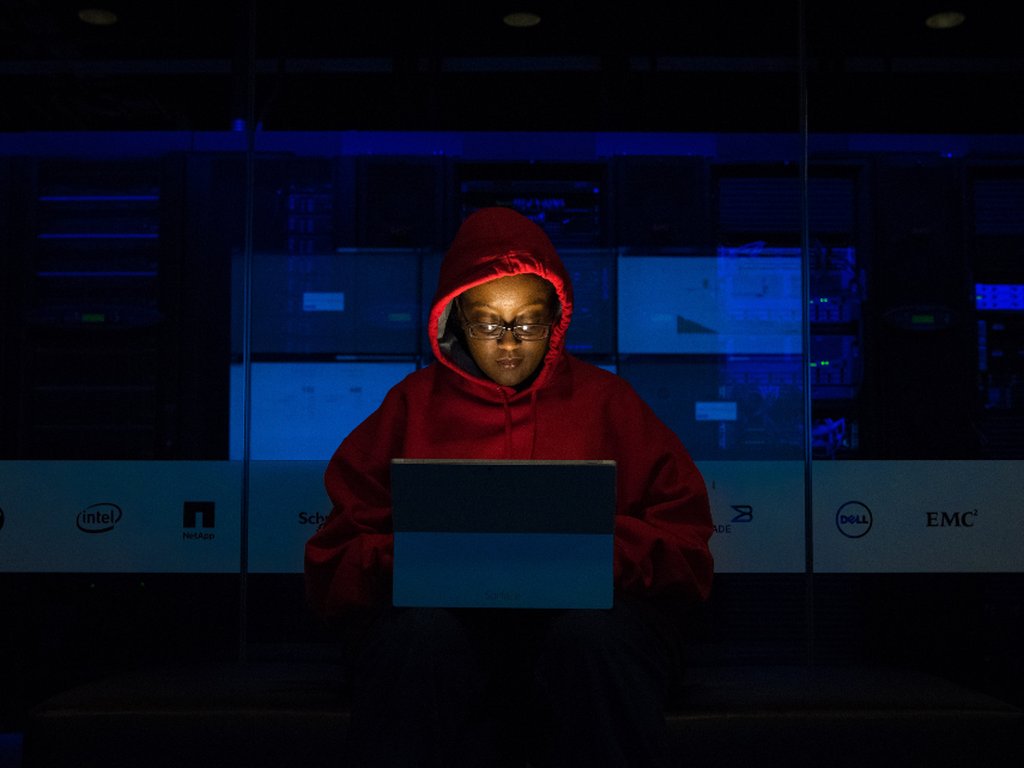 A person in a red hood working on a laptop in a dark room.