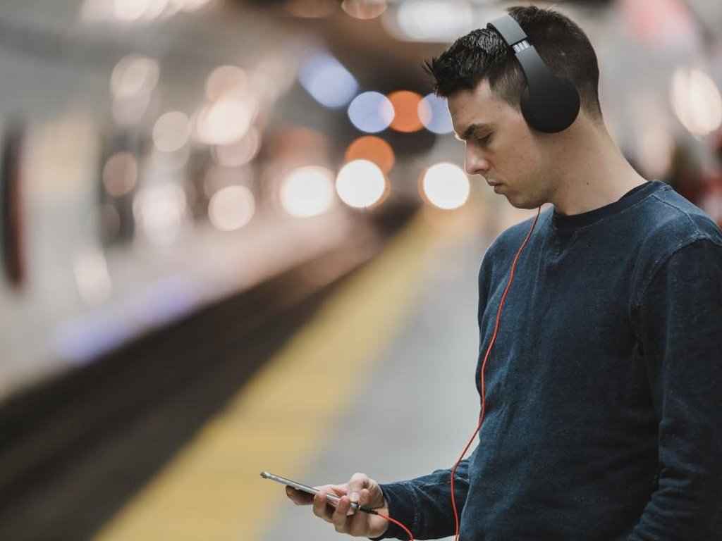 A young man with headphones looking at his phone on an underground platform.