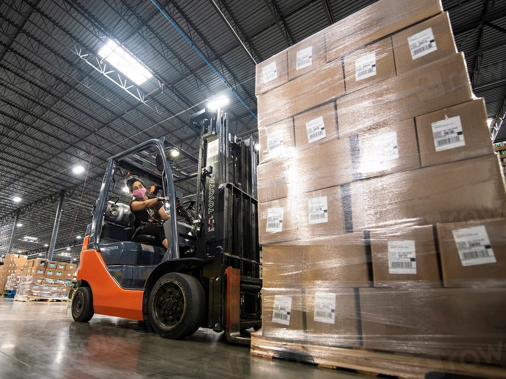 A forklift driver moving a tall stack of boxes in a large warehouse.