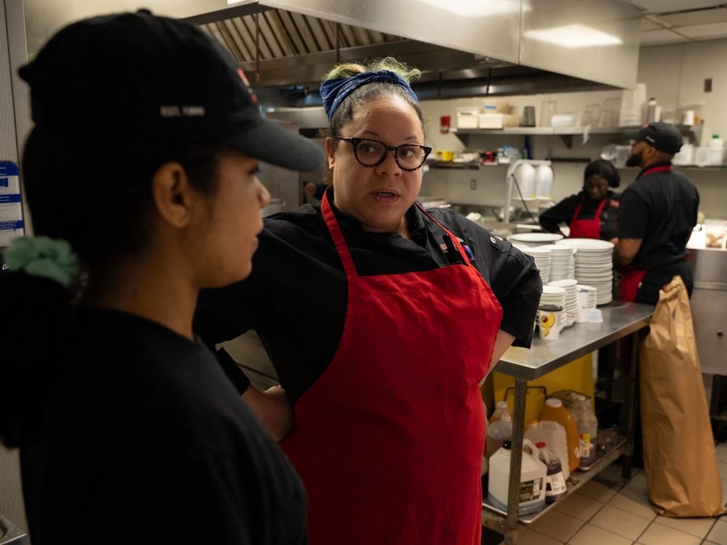 A chef in a red apron talking to a colleague in a busy restaurant kitchen.