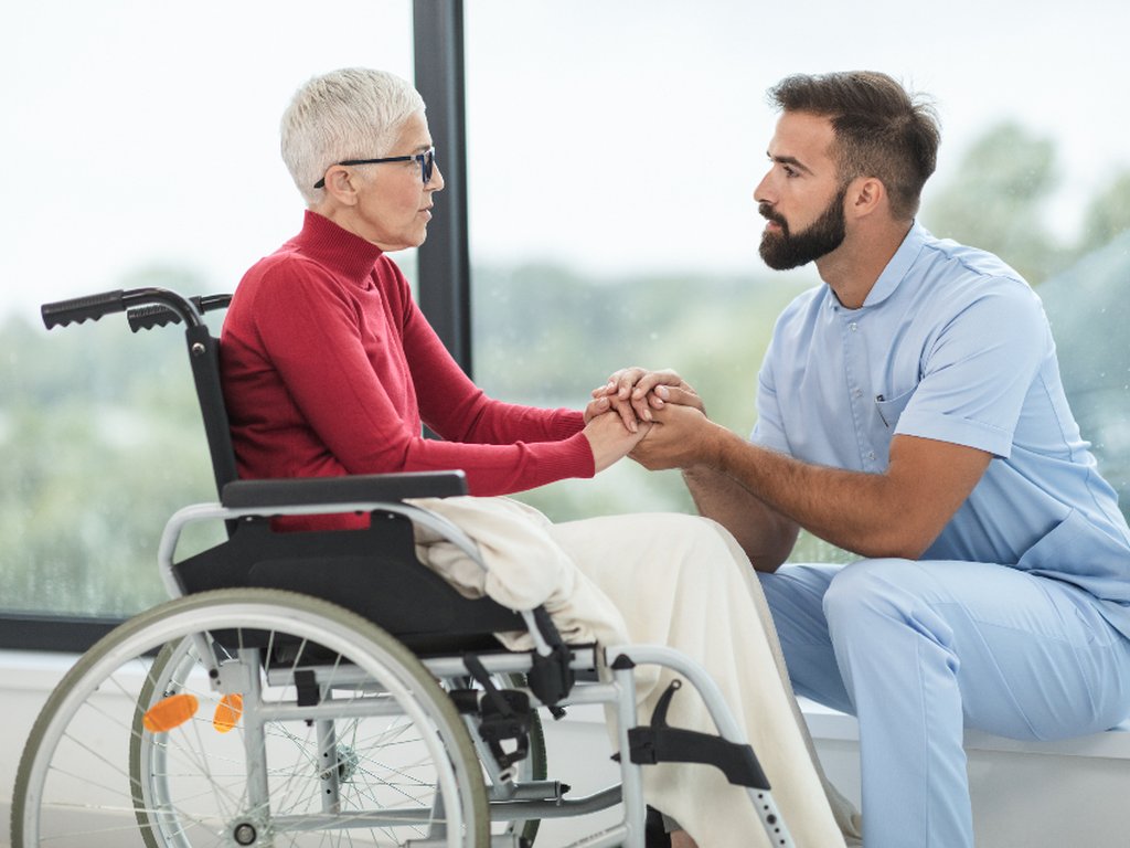 A care worker holding hands and talking with an older woman who uses a wheelchair.