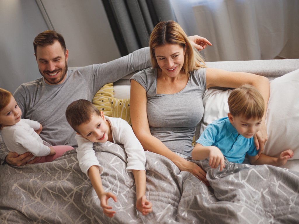 Two parents in bed early in the morning, with their three young children climbing on the bed.
