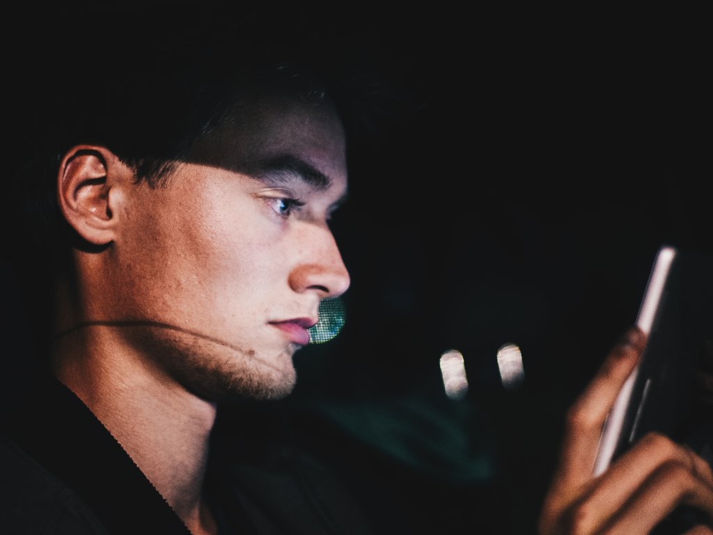 A young man looking at his phone in the dark, his face lit up by the screen.