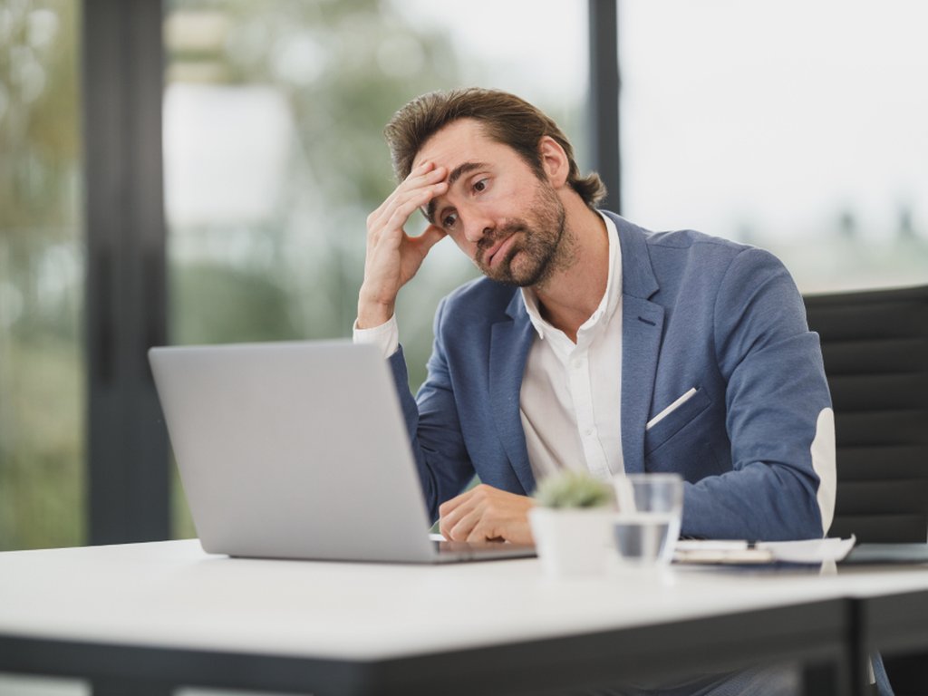 A man at a laptop in an office, holding his head and looking exhausted.